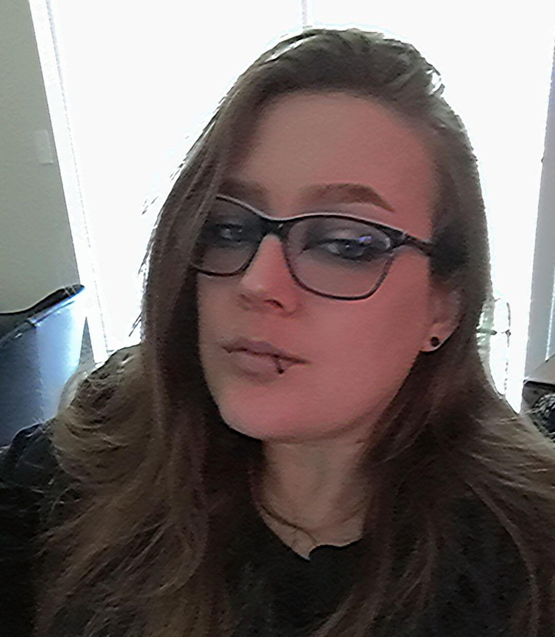 Zab has long blonde straight hair and is wearing purple glasses, black lip ring, earrings and a black shirt.